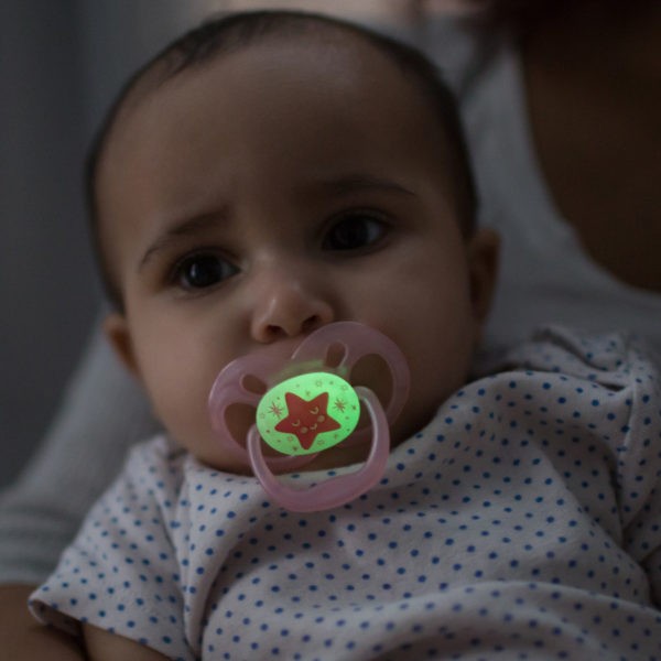 Baby in mother's arms in dark room with glow-in-the-dark pacifier in mouth