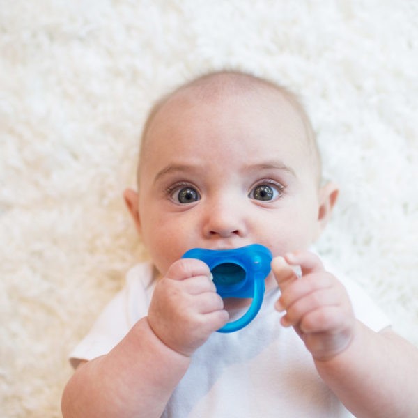 Baby looking up at camera, laying on floor, with blue pacifier in mouth