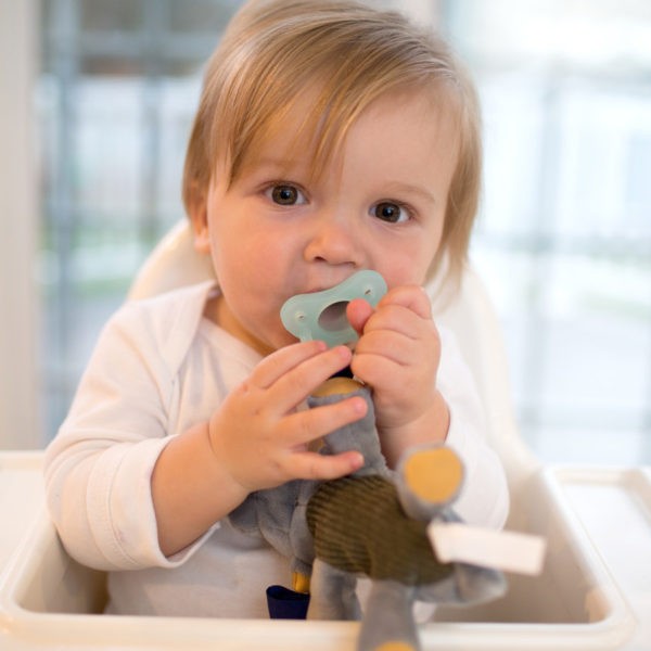 Baby sitting in highchair with triceratops lovey pacifier and teether holder