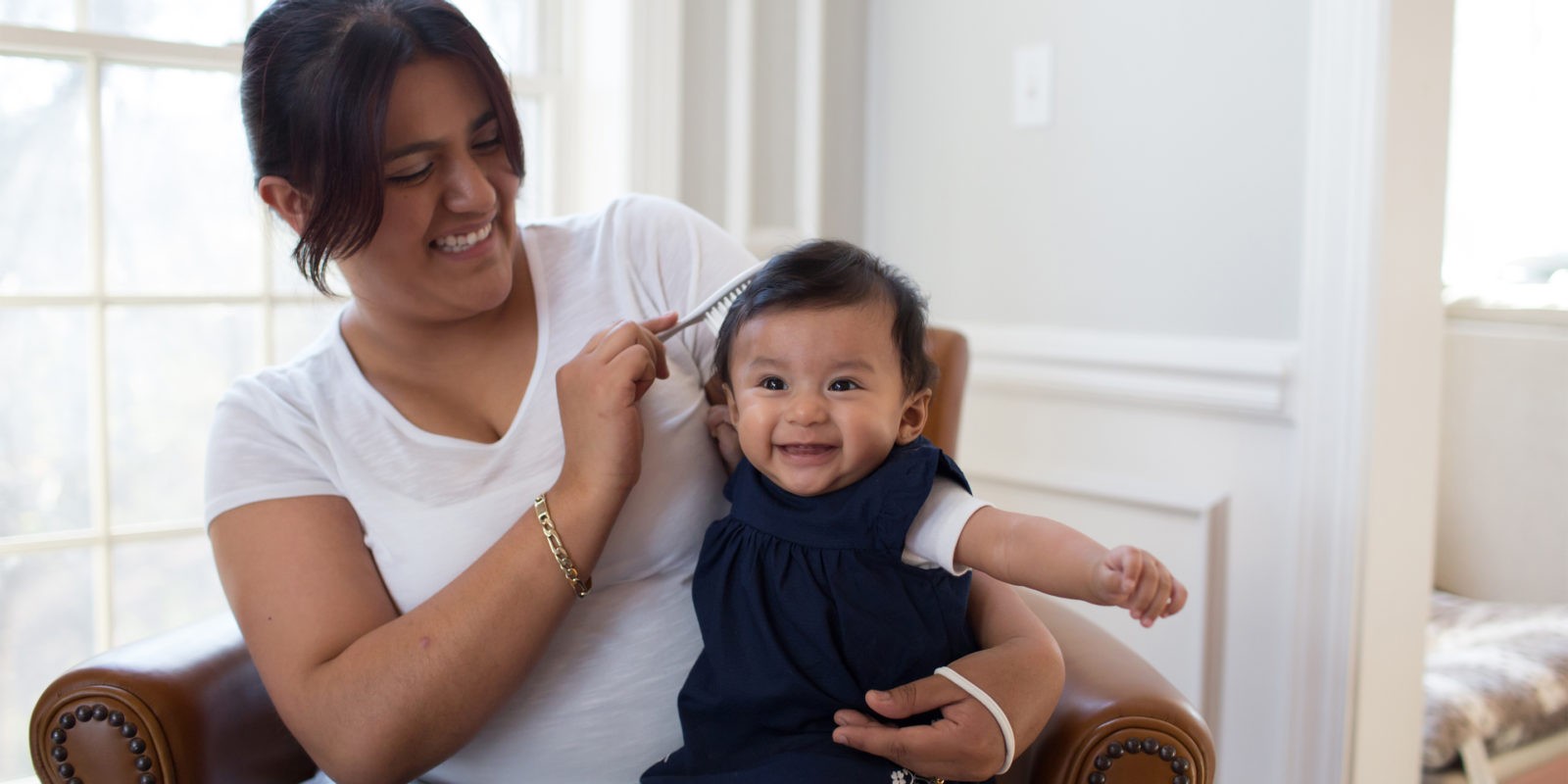 Baby in mother's lap sitting on chair, smiling while hair is being brushed