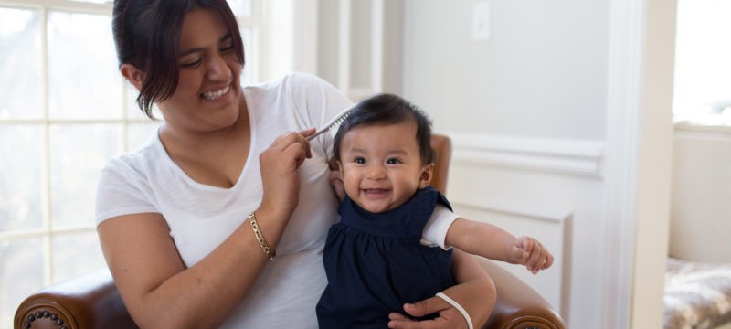 Baby in mother's lap sitting on chair, smiling while hair is being brushed