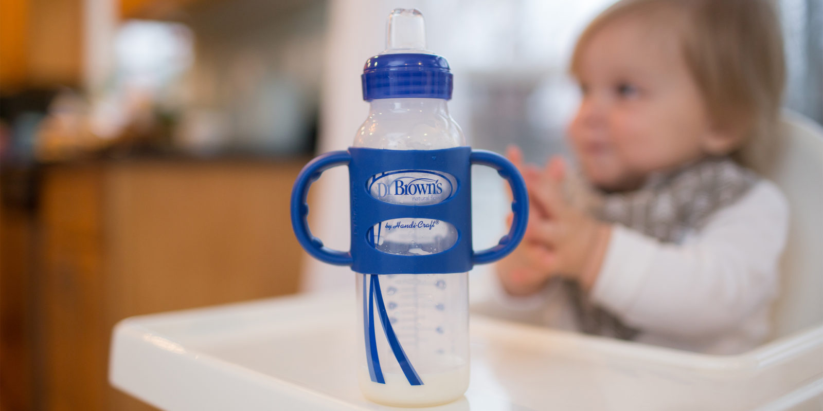 Sippy bottle with silicone handles sitting on highchair table with baby in background