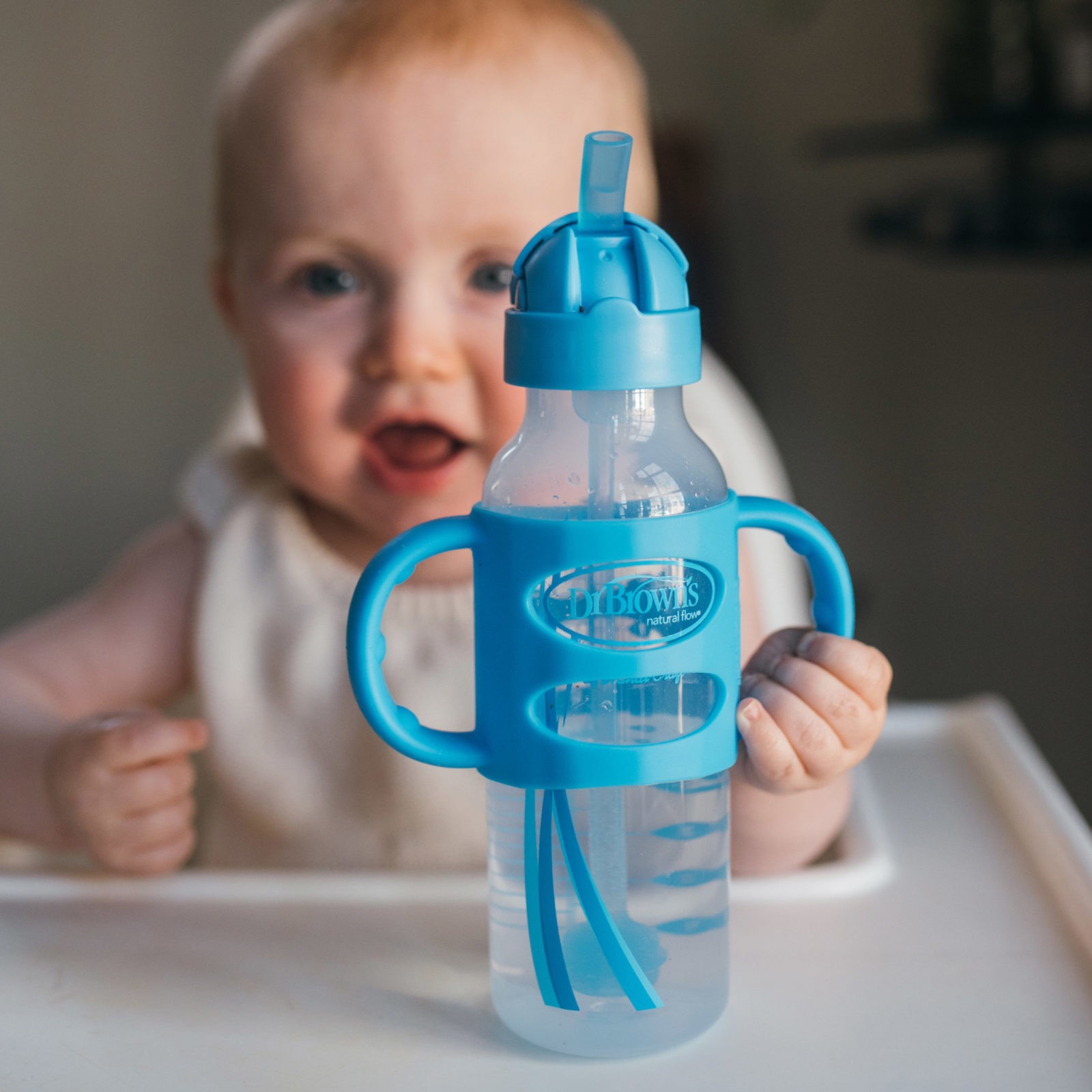 Baby in high chair holding silicone handle on blue sippy straw bottle