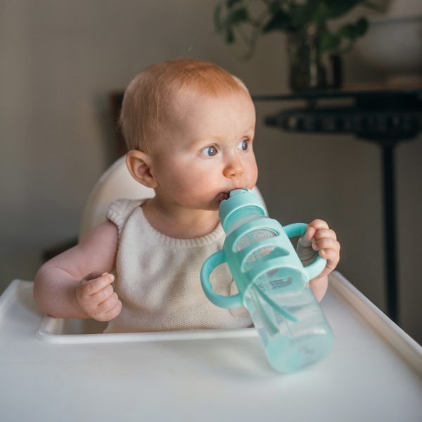 Baby in high chair drinking from green sippy straw bottle