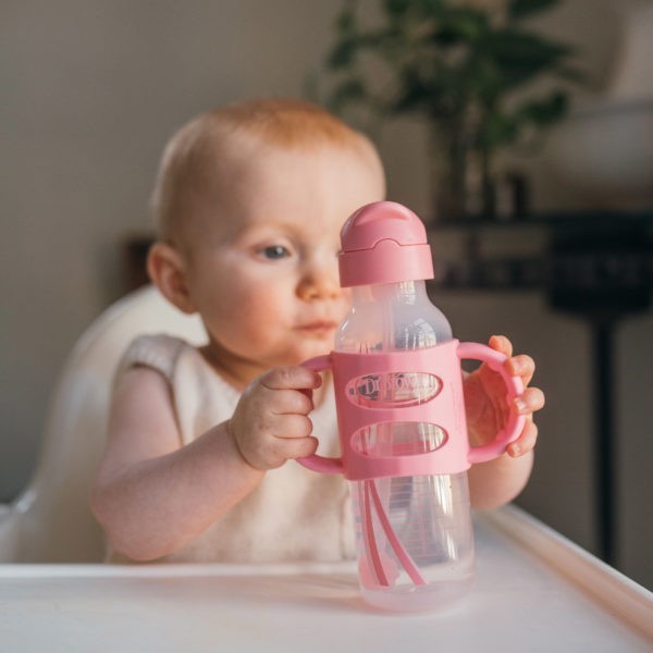 Baby in high chair holding handle on pink sippy straw bottle